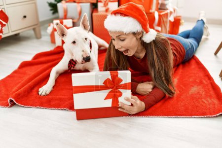Foto de Joven mujer caucásica unboxing regalo acostado con perro por árbol de Navidad en casa - Imagen libre de derechos