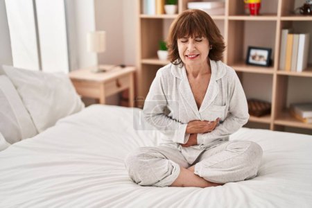 Téléchargez les photos : Femme d'âge moyen souffrant de maux d'estomac assis sur le lit dans la chambre - en image libre de droit