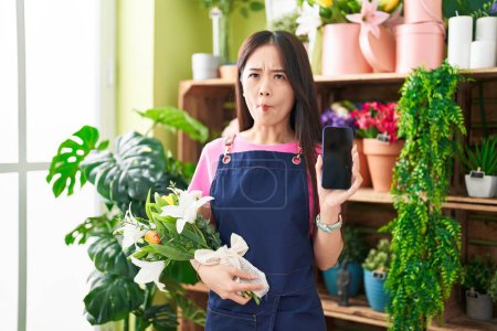 Téléchargez les photos : Jeune femme chinoise travaillant à la boutique de fleuristes montrant écran smartphone faire visage de poisson avec la bouche et les yeux plissés, fou et comique. - en image libre de droit