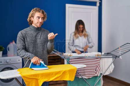 Photo for Couple ironing clothes at laundry room pointing thumb up to the side smiling happy with open mouth - Royalty Free Image
