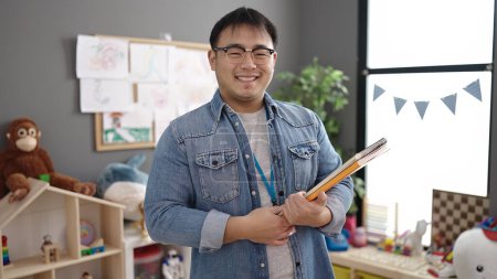 Téléchargez les photos : Jeune enseignant chinois souriant confiant tenant des livres à la maternelle - en image libre de droit