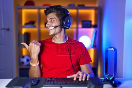 Foto de Joven hispano jugando videojuegos sonriendo con la cara feliz mirando y apuntando hacia un lado con el pulgar hacia arriba. - Imagen libre de derechos