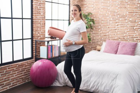 Téléchargez les photos : Jeune femme enceinte tenant tapis de pilate dans la chambre à coucher souriant avec un sourire heureux et cool sur le visage. montrant les dents. - en image libre de droit