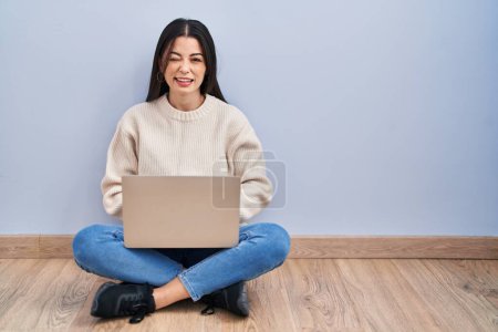 Téléchargez les photos : Jeune femme utilisant un ordinateur portable assis sur le sol à la maison clin d'oeil en regardant la caméra avec une expression sexy, gai et heureux visage. - en image libre de droit