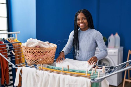 Foto de Mujer afroamericana sonriendo confiada colgando ropa en tendedero en la lavandería - Imagen libre de derechos