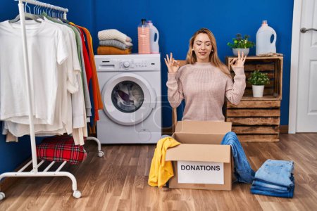 Photo for Hispanic woman putting clothes in donation box relax and smiling with eyes closed doing meditation gesture with fingers. yoga concept. - Royalty Free Image