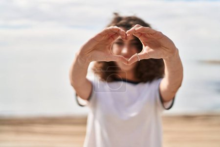 Téléchargez les photos : Jeune femme hispanique souriante confiante faisant geste de cœur avec les mains au bord de la mer - en image libre de droit