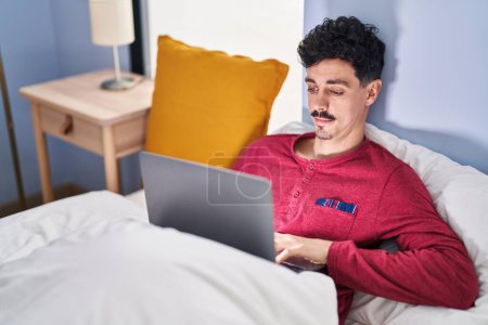 Foto de Hombre caucásico joven usando el ordenador portátil sentado en la cama en el dormitorio - Imagen libre de derechos