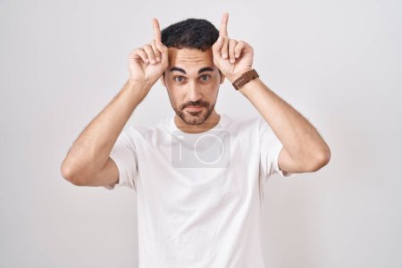 Téléchargez les photos : Bel homme hispanique debout sur fond blanc faisant geste drôle avec doigt sur tête comme cornes de taureau - en image libre de droit
