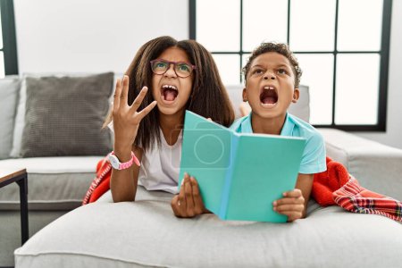 Photo for Two siblings lying on the sofa reading a book crazy and mad shouting and yelling with aggressive expression and arms raised. frustration concept. - Royalty Free Image