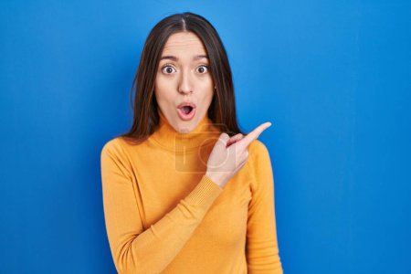 Photo for Young brunette woman standing over blue background surprised pointing with finger to the side, open mouth amazed expression. - Royalty Free Image