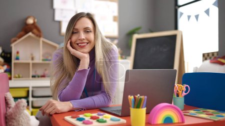 Téléchargez les photos : Jeune femme blonde prof de maternelle en utilisant un ordinateur portable assis sur la table à la maternelle - en image libre de droit