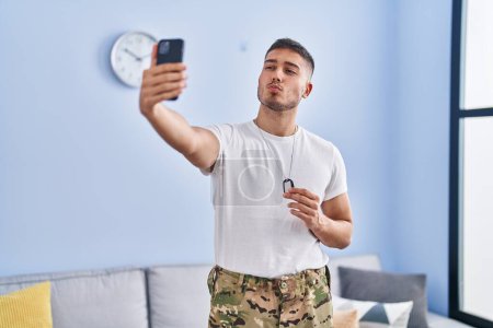 Téléchargez les photos : Jeune homme hispanique portant un uniforme de camouflage de l'armée prenant selfie à la maison en regardant la caméra souffler un baiser étant belle et sexy. expression d'amour. - en image libre de droit