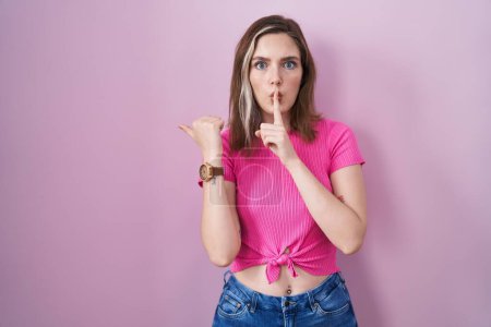 Photo for Blonde caucasian woman standing over pink background asking to be quiet with finger on lips pointing with hand to the side. silence and secret concept. - Royalty Free Image
