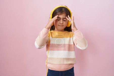Photo for Little hispanic girl listening to music using headphones with hand on head, headache because stress. suffering migraine. - Royalty Free Image