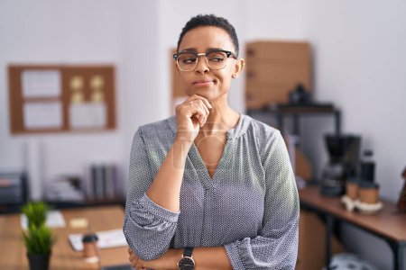 Téléchargez les photos : Femme afro-américaine travaillant au bureau portant des lunettes regardant avec confiance la caméra avec le sourire avec les bras croisés et la main levée sur le menton. penser positivement. - en image libre de droit