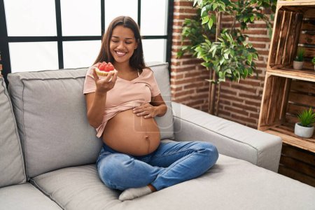 Photo for Young latin woman pregnant eating cake sitting on sofa at home - Royalty Free Image