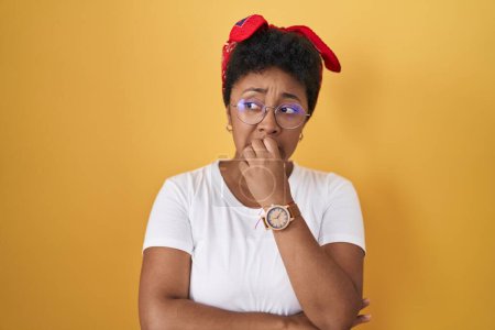 Foto de Mujer afroamericana joven de pie sobre fondo amarillo mirando estresada y nerviosa con las manos en la boca mordiendo las uñas. problema de ansiedad. - Imagen libre de derechos
