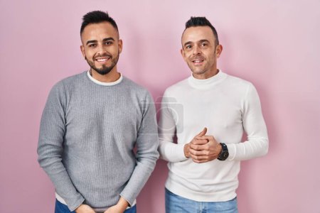 Téléchargez les photos : Couple homosexuel debout sur fond rose avec les mains jointes et les doigts croisés souriant détendu et joyeux. succès et optimisme - en image libre de droit