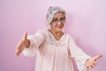 Foto de Mujer de mediana edad con el pelo gris de pie sobre fondo rosa mirando a la cámara sonriendo con los brazos abiertos para el abrazo. expresión alegre abrazando la felicidad. - Imagen libre de derechos