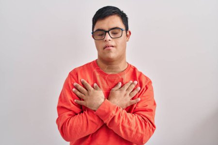 Photo for Young hispanic man with down syndrome standing over white background smiling with hands on chest, eyes closed with grateful gesture on face. health concept. - Royalty Free Image