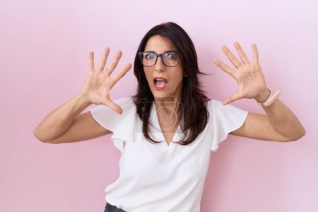 Photo for Middle age hispanic woman wearing casual white t shirt and glasses afraid and terrified with fear expression stop gesture with hands, shouting in shock. panic concept. - Royalty Free Image