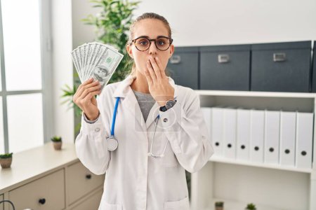 Photo for Young doctor woman holding dollars banknotes covering mouth with hand, shocked and afraid for mistake. surprised expression - Royalty Free Image