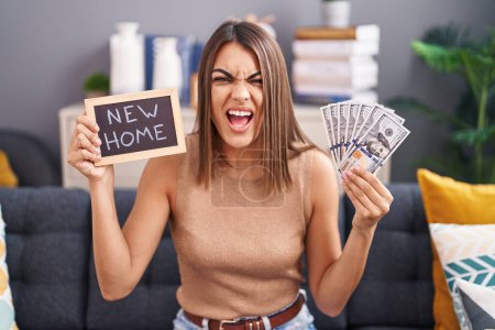 Photo for Young hispanic woman holding blackboard with new home text and dollars angry and mad screaming frustrated and furious, shouting with anger. rage and aggressive concept. - Royalty Free Image
