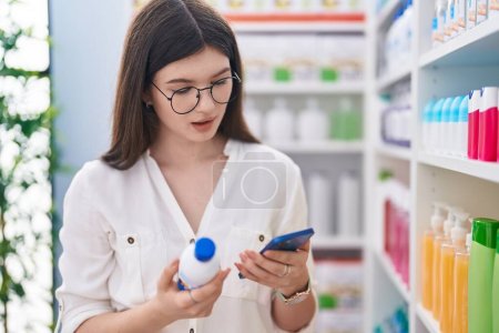 Téléchargez les photos : Jeune cliente caucasienne utilisant un smartphone tenant un flacon de médicament à la pharmacie - en image libre de droit