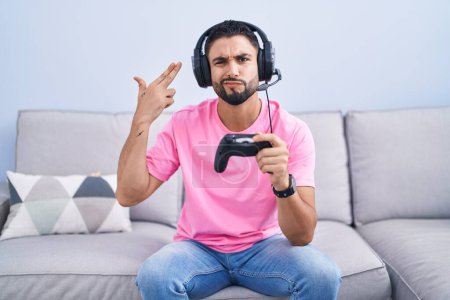 Foto de Joven hispano jugando videojuego sosteniendo controlador sentado en el sofá disparando y matándose apuntando de la mano y los dedos a la cabeza como arma, gesto suicida. - Imagen libre de derechos