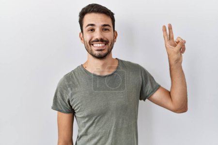 Téléchargez les photos : Jeune homme hispanique à la barbe portant un t-shirt décontracté sur fond blanc souriant avec un clin d'œil heureux à la caméra faisant signe de victoire. numéro deux. - en image libre de droit