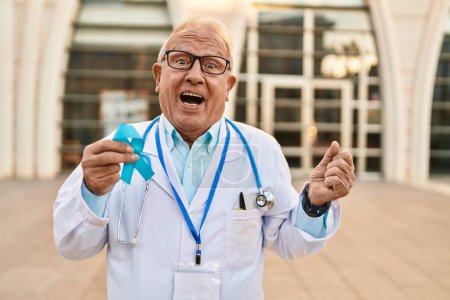 Téléchargez les photos : Médecin sénior aux cheveux gris tenant un ruban bleu criant fier, célébrant la victoire et le succès très excité avec les bras levés - en image libre de droit