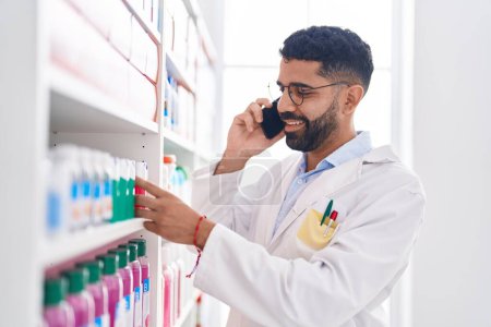 Téléchargez les photos : Jeune homme arabe pharmacien tenant bouteille de dentifrice parler sur smartphone à la pharmacie - en image libre de droit