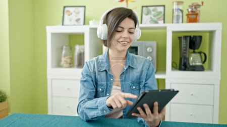 Foto de Mujer joven caucásica usando touchpad y auriculares sentados en la mesa en casa - Imagen libre de derechos