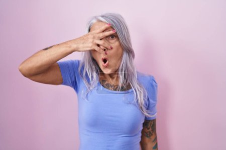 Foto de Mujer de mediana edad con tatuajes de pie sobre fondo rosa asomándose en shock cubriendo la cara y los ojos con la mano, mirando a través de los dedos con expresión avergonzada. - Imagen libre de derechos