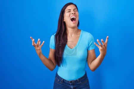 Photo for Young hispanic woman standing over blue background crazy and mad shouting and yelling with aggressive expression and arms raised. frustration concept. - Royalty Free Image
