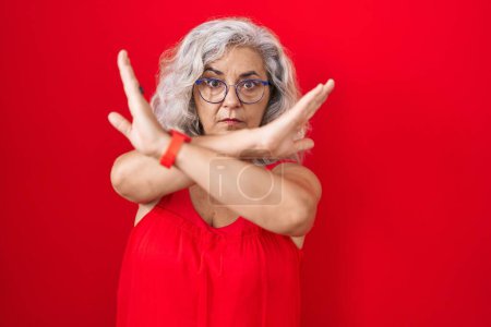 Téléchargez les photos : Femme d'âge moyen avec les cheveux gris debout sur fond rouge expression de rejet croisant les bras faisant signe négatif, visage en colère - en image libre de droit
