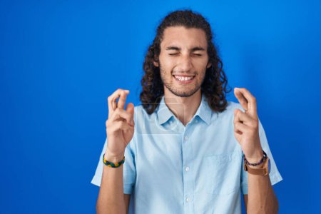 Téléchargez les photos : Jeune homme hispanique debout sur fond bleu geste doigt croisé souriant avec espoir et les yeux fermés. chance et concept superstitieux. - en image libre de droit
