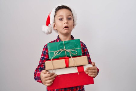 Foto de Pequeño niño hispano con sombrero de navidad sosteniendo regalos en la cara de choque, mirando escéptico y sarcástico, sorprendido con la boca abierta - Imagen libre de derechos