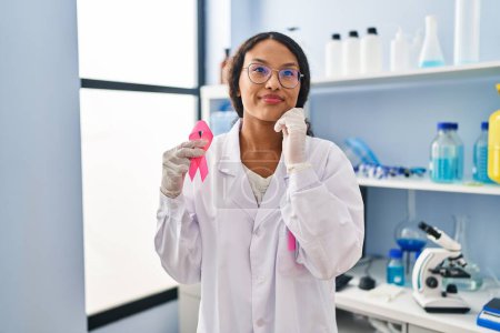 Téléchargez les photos : Jeune médecin hispanique femme travaillant au laboratoire scientifique tenant ruban rose visage sérieux penser à la question avec la main sur le menton, réfléchi sur l'idée déroutante - en image libre de droit