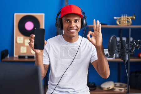 Foto de Joven hombre hispano escuchando música usando auriculares que muestran la pantalla del teléfono inteligente haciendo signo bien con los dedos, sonriendo gesto amistoso excelente símbolo - Imagen libre de derechos