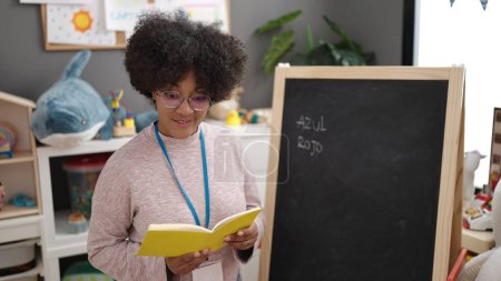 Foto de Joven afroamericana mujer preescolar profesor lectura libro en el jardín de infantes - Imagen libre de derechos