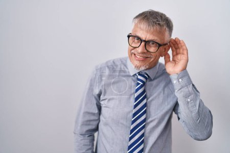 Téléchargez les photos : Homme d'affaires hispanique aux cheveux gris portant des lunettes souriant main sur oreille en écoutant une rumeur ou des commérages. concept de surdité. - en image libre de droit