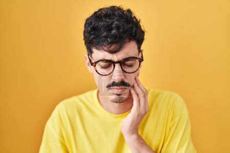 Téléchargez les photos : Homme hispanique portant des lunettes debout sur fond jaune touchant la bouche avec la main avec une expression douloureuse à cause de maux de dents ou de maladies dentaires sur les dents. dentiste - en image libre de droit