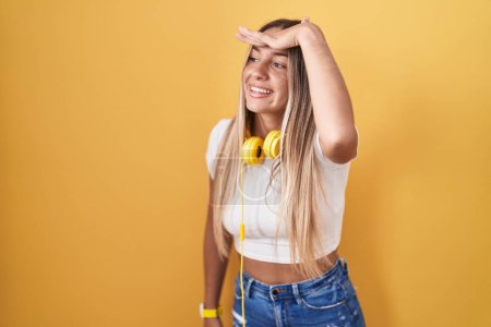 Foto de Mujer rubia joven de pie sobre fondo amarillo con auriculares muy felices y sonrientes mirando lejos con la mano sobre la cabeza. concepto de búsqueda. - Imagen libre de derechos