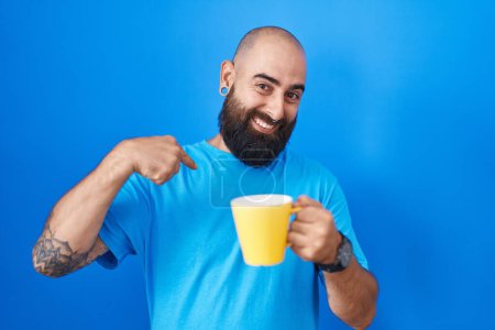 Foto de Joven hombre hispano con barba y tatuajes bebiendo una taza de café mirando confiado con sonrisa en la cara, señalándose con los dedos orgullosos y felices. - Imagen libre de derechos