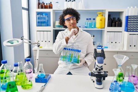 Photo for Hispanic man with curly hair working at scientist laboratory serious face thinking about question with hand on chin, thoughtful about confusing idea - Royalty Free Image