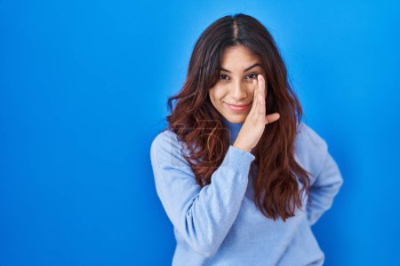 Photo for Hispanic young woman standing over blue background hand on mouth telling secret rumor, whispering malicious talk conversation - Royalty Free Image