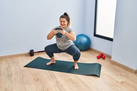 Photo for Young beautiful plus size woman using kettlebell training legs exercise at sport center - Royalty Free Image