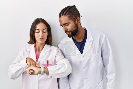 Téléchargez les photos : Jeunes médecins hispaniques debout sur fond blanc vérifiant l'heure sur la montre-bracelet, détendu et confiant - en image libre de droit